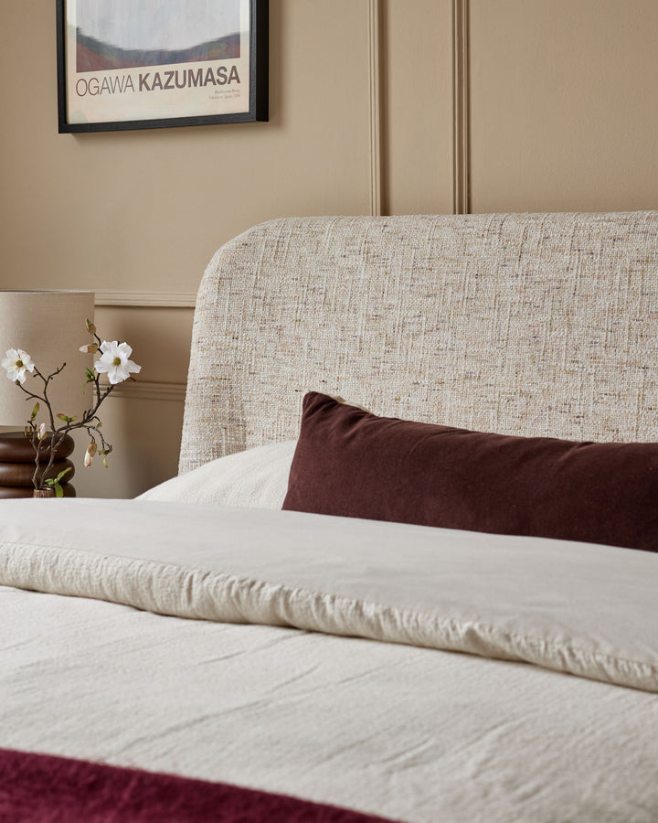 Deven King-Size Ottoman Bed in natural fabric, showing neutral upholstered headboard with cream bedding and burgundy accent pillow.