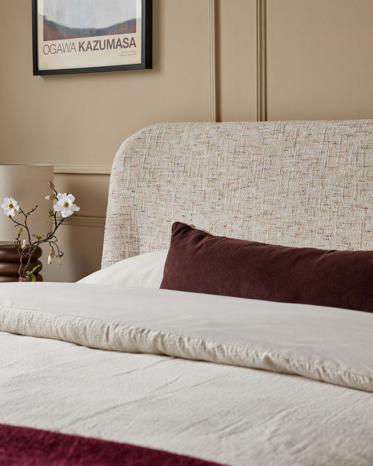 Deven King-Size Ottoman Bed in natural fabric, showing neutral upholstered headboard with cream bedding and burgundy accent pillow.
