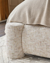 Deven king-size ottoman bed in natural fabric, close-up of curved cylindrical foot detail and herringbone pattern cushion.