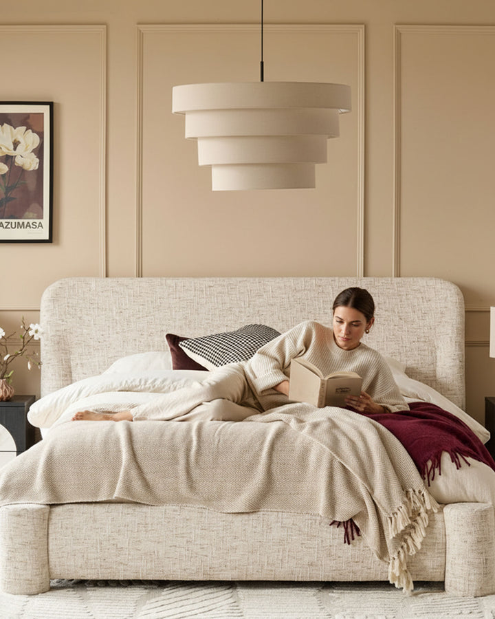 Deven King-Size Ottoman Bed in natural fabric with rounded headboard, shown in a styled bedroom with woman reading.