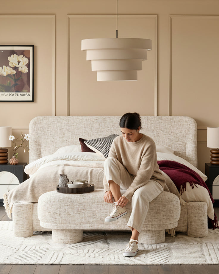 Deven Bench in natural fabric, rounded cylindrical shape with textured upholstery, shown at foot of bed in bedroom setting.