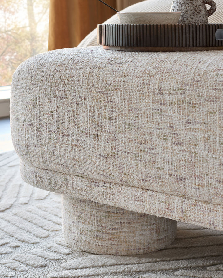 Deven bench in natural fabric, round cylindrical shape with textured beige upholstery and curved seating surface.