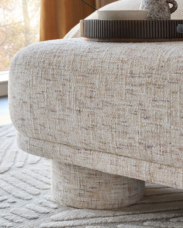 Deven bench in natural fabric, round cylindrical shape with textured beige upholstery and curved seating surface.