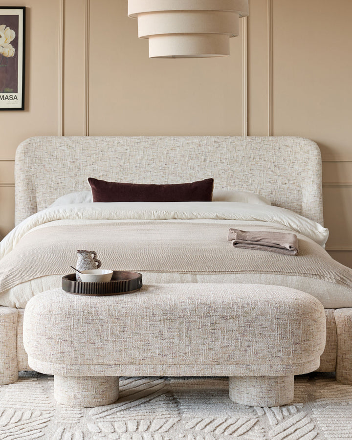 Deven Bench in natural fabric with rounded edges, positioned at foot of bed in neutral bedroom setting.