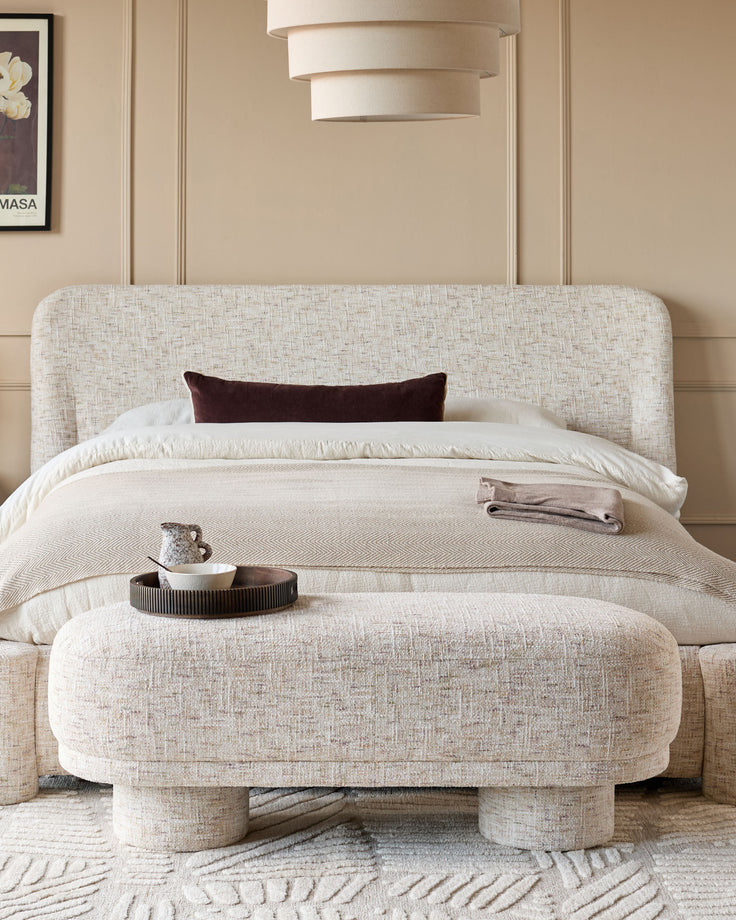 Deven Bench in natural fabric with rounded edges, positioned at foot of bed in neutral bedroom setting.