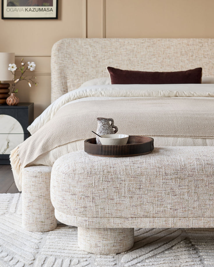 Deven bench in natural fabric with cylindrical legs, styled at the foot of a bed in a contemporary bedroom setting.