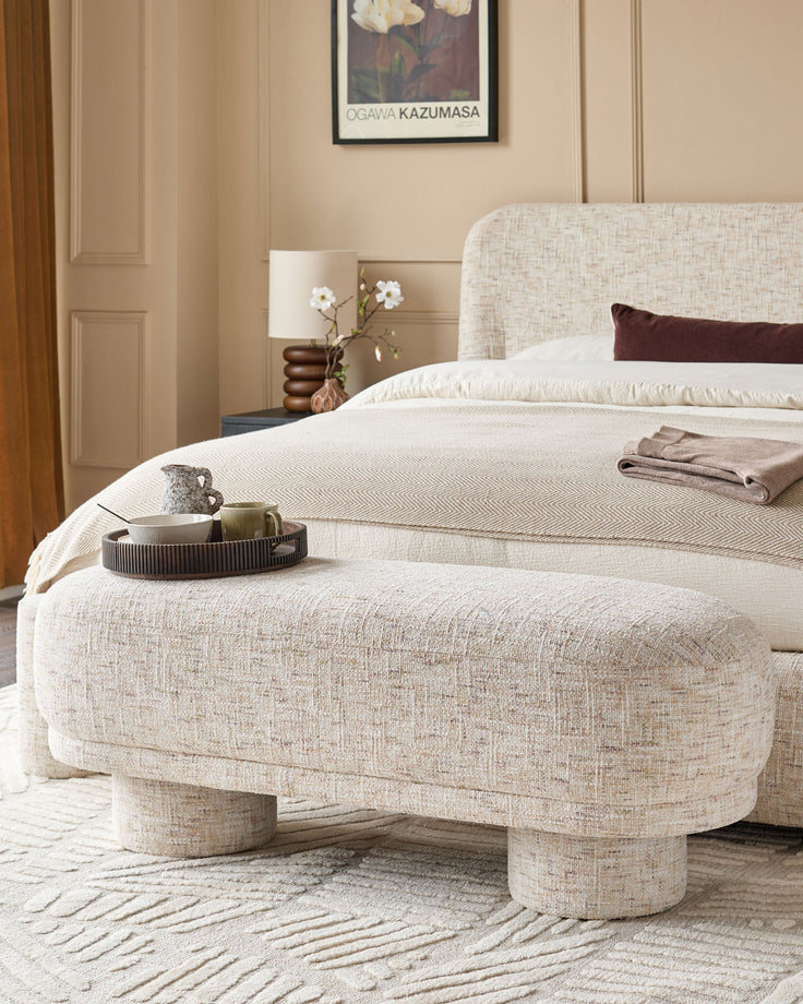 Deven bench in natural fabric with curved, rounded silhouette and cylindrical legs, positioned at foot of bed.