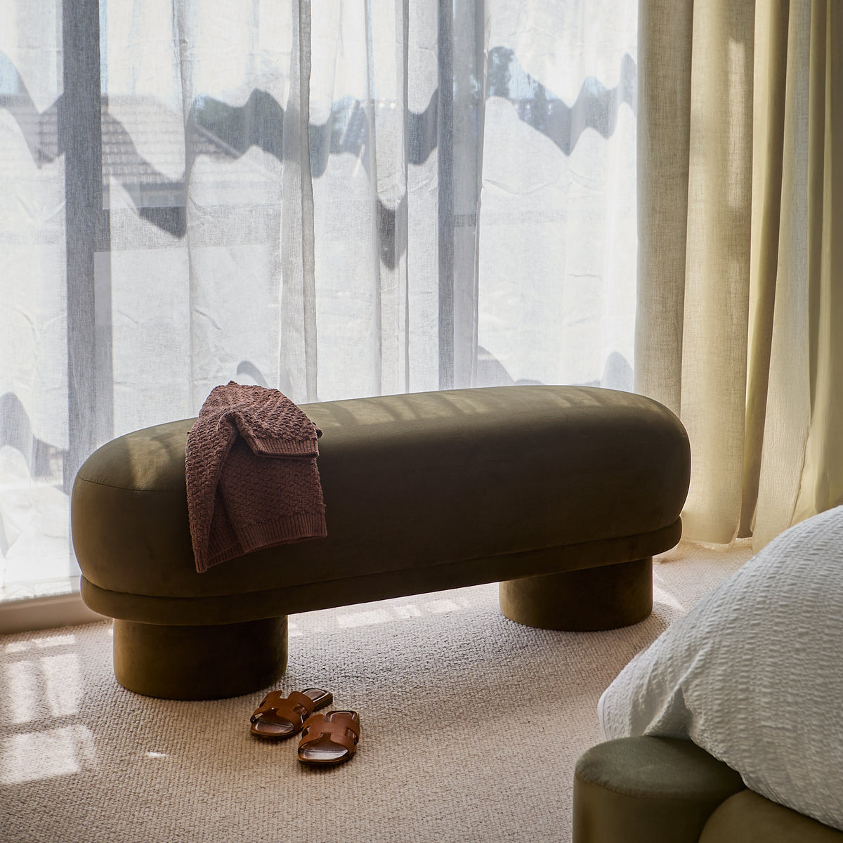Deven bench in olive green velvet with cylindrical legs, styled in a bright bedroom with sheer curtains and warm lighting.