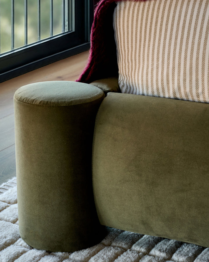Olive green velvet ottoman with cylindrical shape, positioned next to striped cushions and window, on neutral rug.