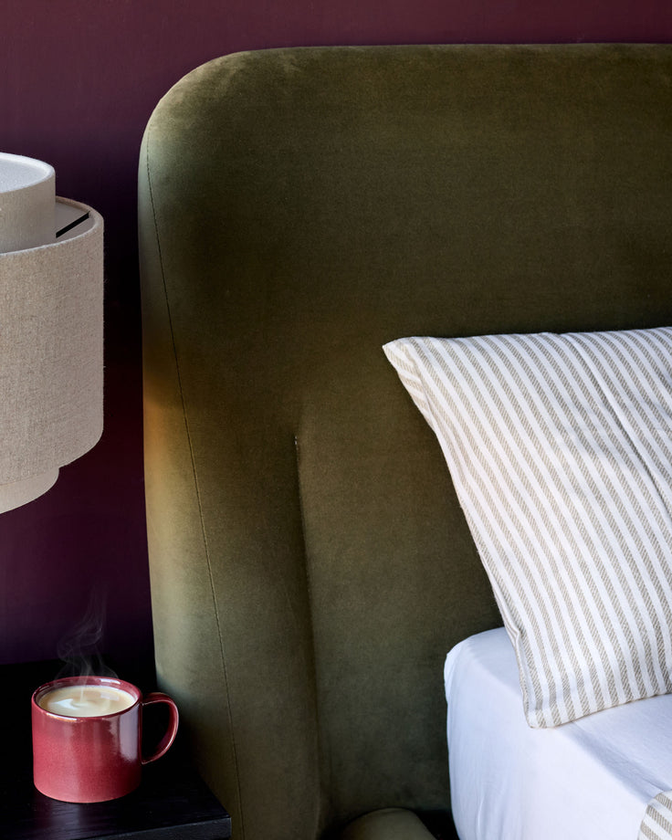 Olive green velvet ottoman bed with rounded headboard, striped pillows, and styled bedding against burgundy wall.