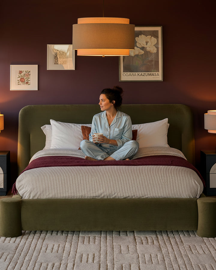 Deven king-size ottoman bed in olive-green velvet with woman sitting on striped bedding in styled bedroom setting.
