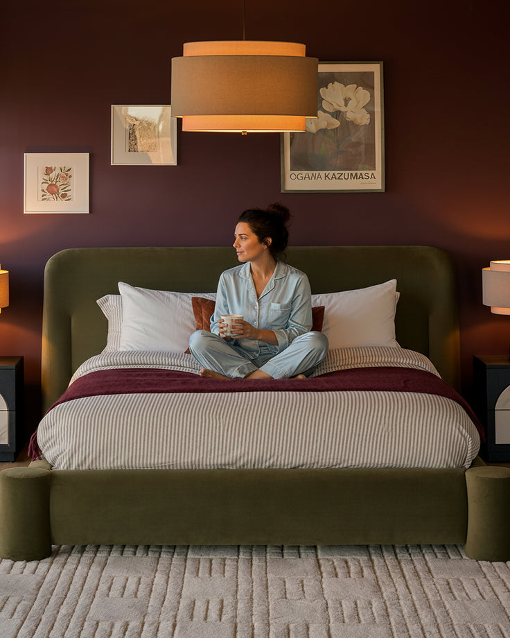 Deven king-size ottoman bed in olive-green velvet with woman sitting on striped bedding in styled bedroom setting.