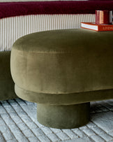 Deven bench in olive green velvet with cylindrical shape, shown in a living room setting on a striped rug.