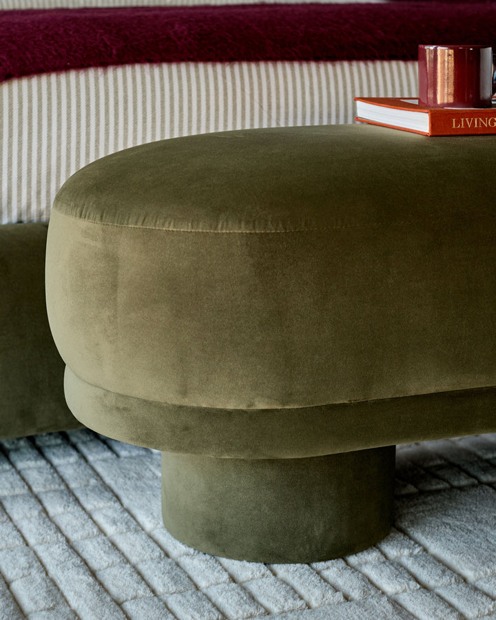 Deven bench in olive green velvet with cylindrical shape, shown in a living room setting on a striped rug.