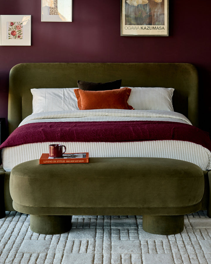Deven Bench in olive green velvet, shown at foot of bed with rounded edges and cylindrical legs.