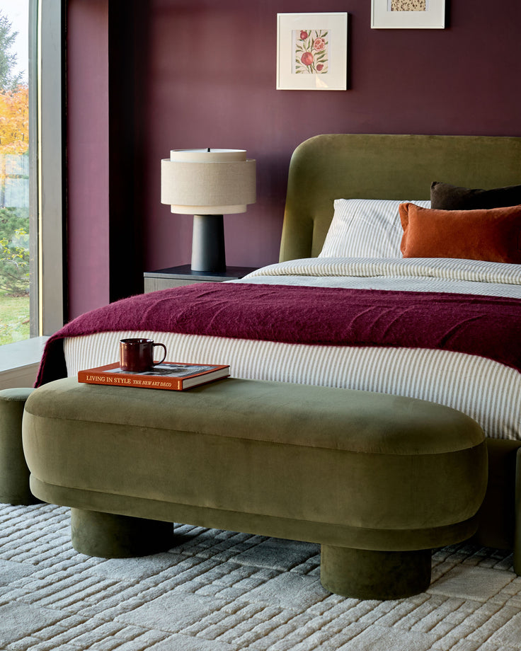 Olive green velvet bench with curved shape and cylindrical legs, positioned at foot of bed in styled bedroom.