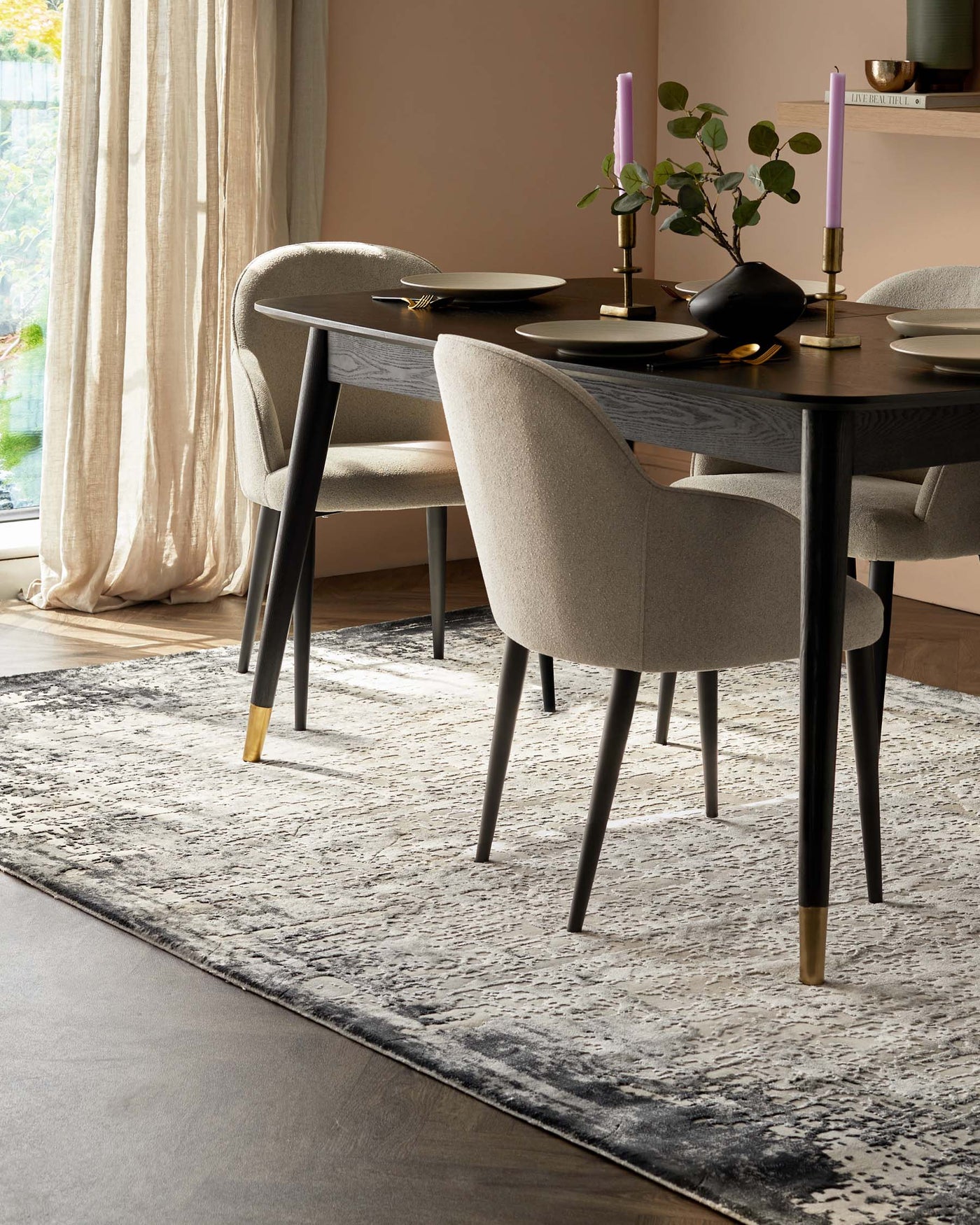 Elegant modern dining area featuring a round, dark wooden table with tapering black legs accented with gold tips. The table is surrounded by four plush, curved-back chairs upholstered in a light, textured fabric, with legs mirroring the table’s black and gold design. The set rests on a large abstract-patterned area rug in shades of grey and cream, anchoring the space on a warm wood floor.