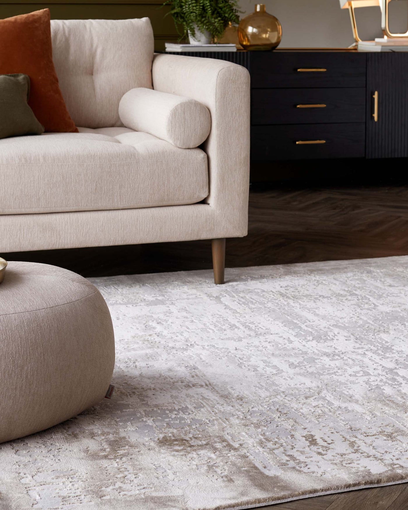 Beige contemporary sofa with a minimalist design featuring round wooden legs and a smooth fabric finish. Paired with a matching round beige ottoman. A sleek dark wood sideboard with gold handles stands in the background atop a textured white area rug with a subtle pattern.
