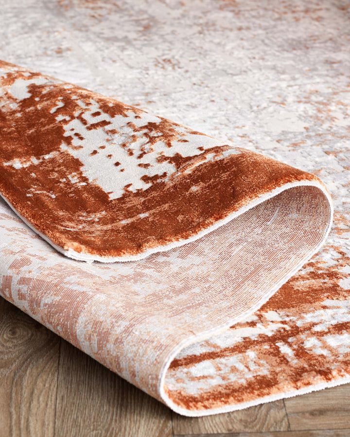 Luxurious textured area rug with a rolled corner, showcasing a plush rust and cream abstract pattern on a wooden floor.