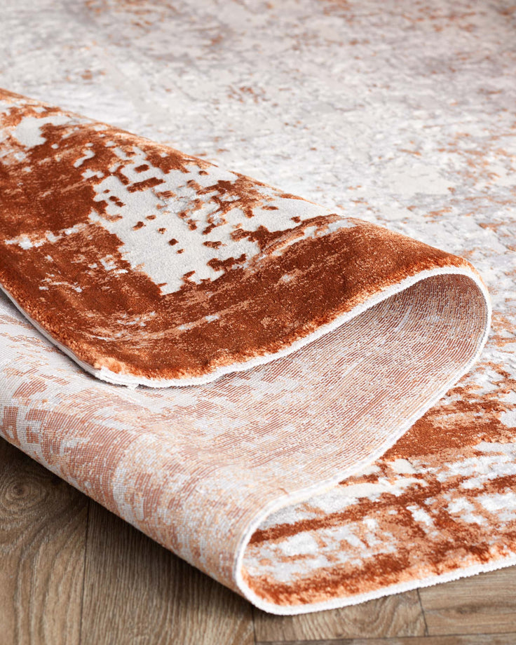 Luxurious textured area rug with a rolled corner, showcasing a plush rust and cream abstract pattern on a wooden floor.