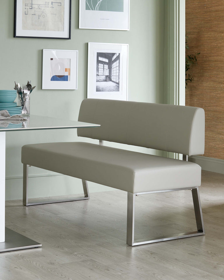 Modern minimalist bench with a tufted, light grey upholstered seat and backrest, featuring sleek, metallic chrome legs in a geometric design. The bench is paired with a simple white rectangular table with a similar chrome leg finish. The room has a contemporary aesthetic with wall art and light hardwood flooring.
