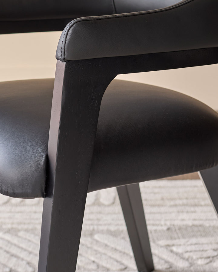 Black leather dining chair with dark wood legs, close-up detail of armrest and frame against light background.