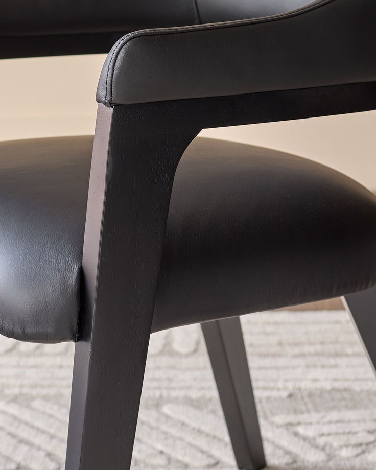 Black leather dining chair with dark wood legs, close-up detail of armrest and frame against light background.
