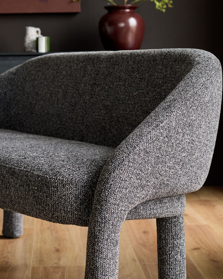 Easton dining bench in dark grey fabric, shown at an angle highlighting its curved backrest and upholstered legs on wood flooring.