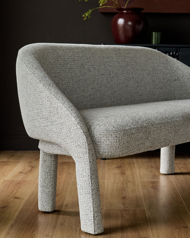 Easton light grey fabric dining bench with curved backrest and cylindrical legs, angled side view on wooden floor.