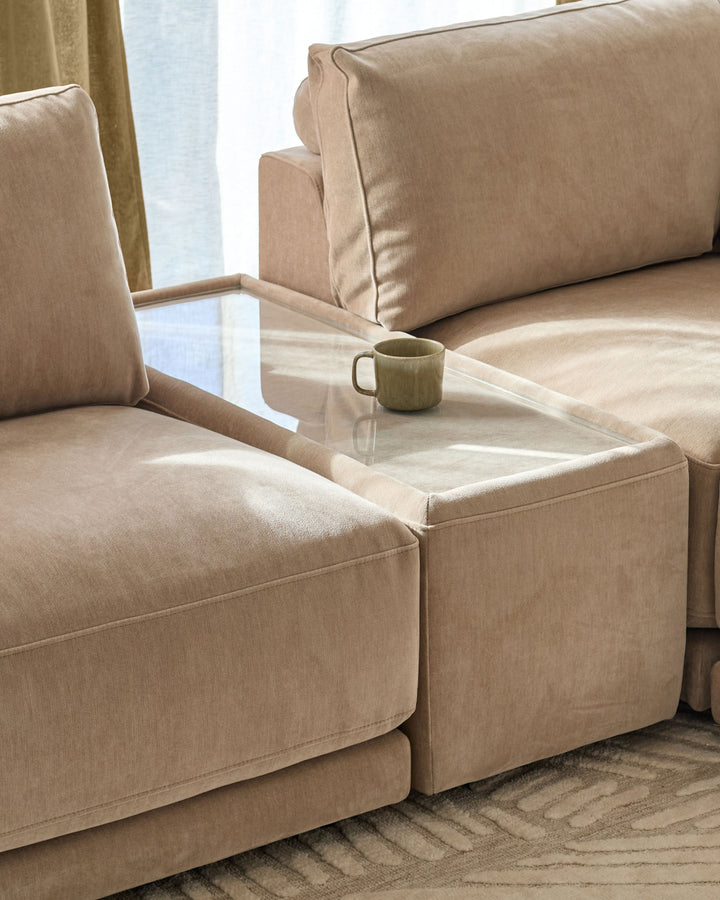Eden side table module with glass top and champagne fabric base, positioned between two beige upholstered seating sections.