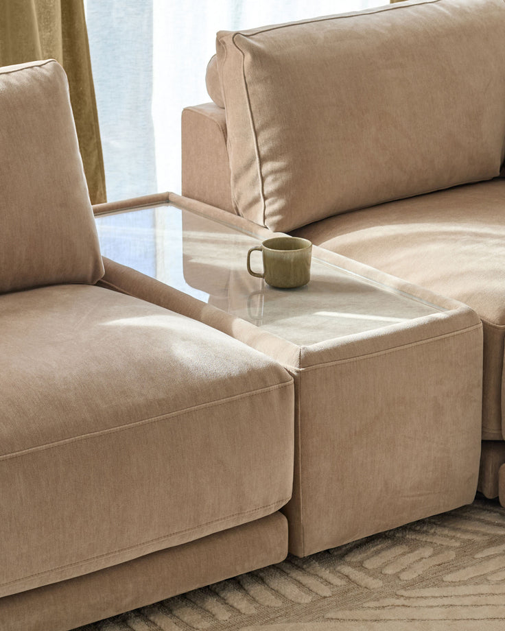 Eden side table module with glass top and champagne fabric base, positioned between two beige upholstered seating sections.