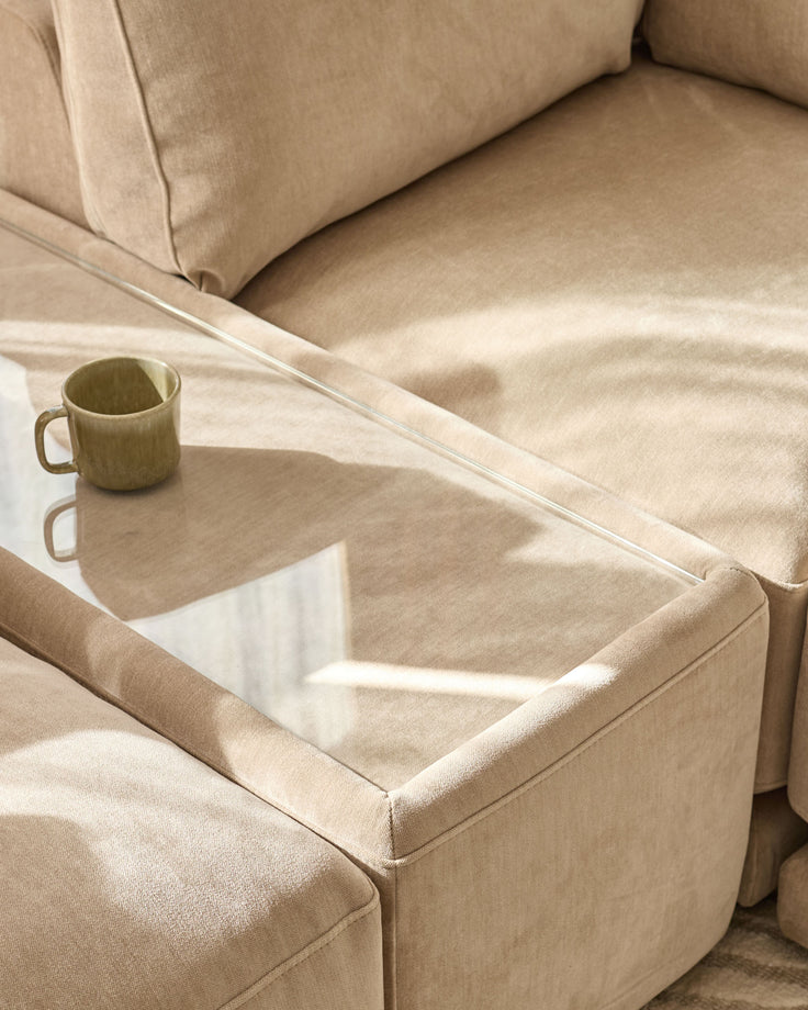 Eden side table module in champagne fabric with glass top, positioned between beige upholstered chairs with warm sunlight.