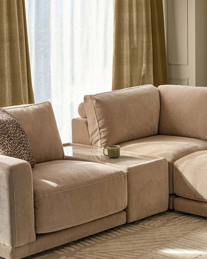 Eden side table module in champagne fabric with glass top, positioned between two tan upholstered armchairs in a bright living room setting.