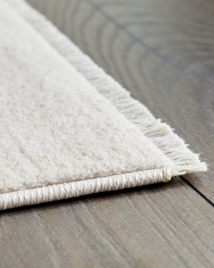 Close-up of an off-white rug with fringed edges, placed on a wood floor.