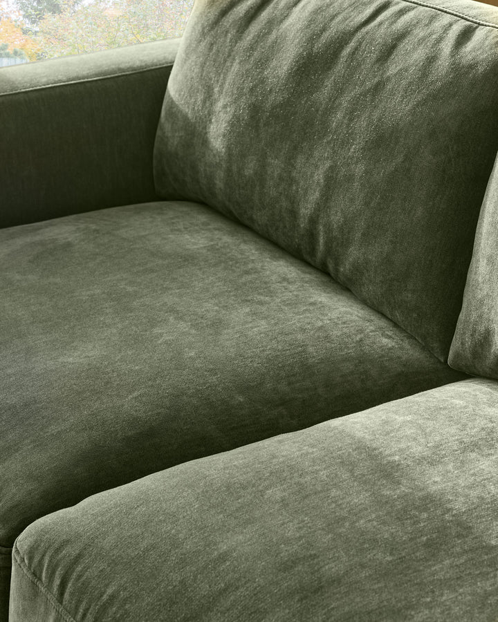 Eden 2-seater sofa in dark green fabric, close-up detail showing seat cushion and backrest with decorative pillow.