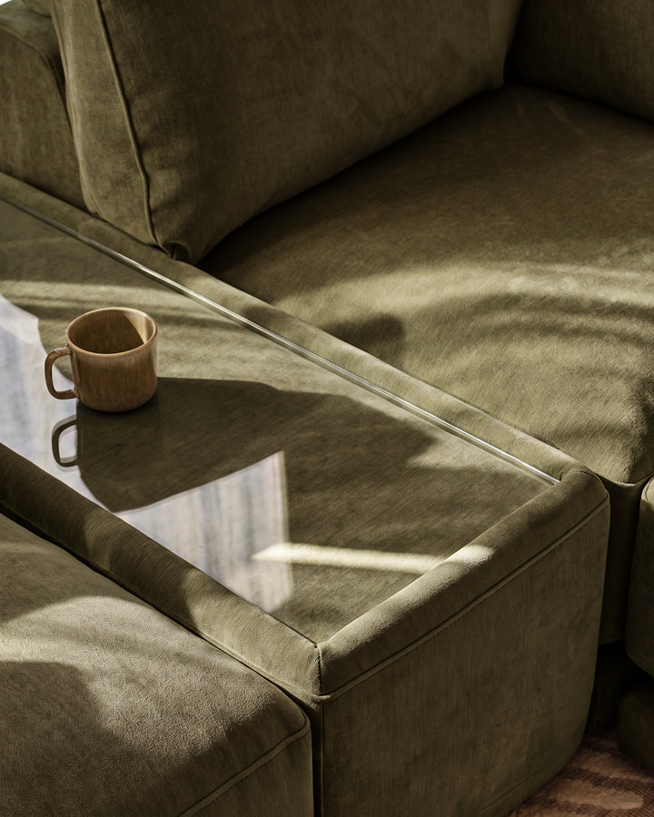 Eden side table module in dark green fabric with glass top, styled with ceramic mug in sunlit setting.