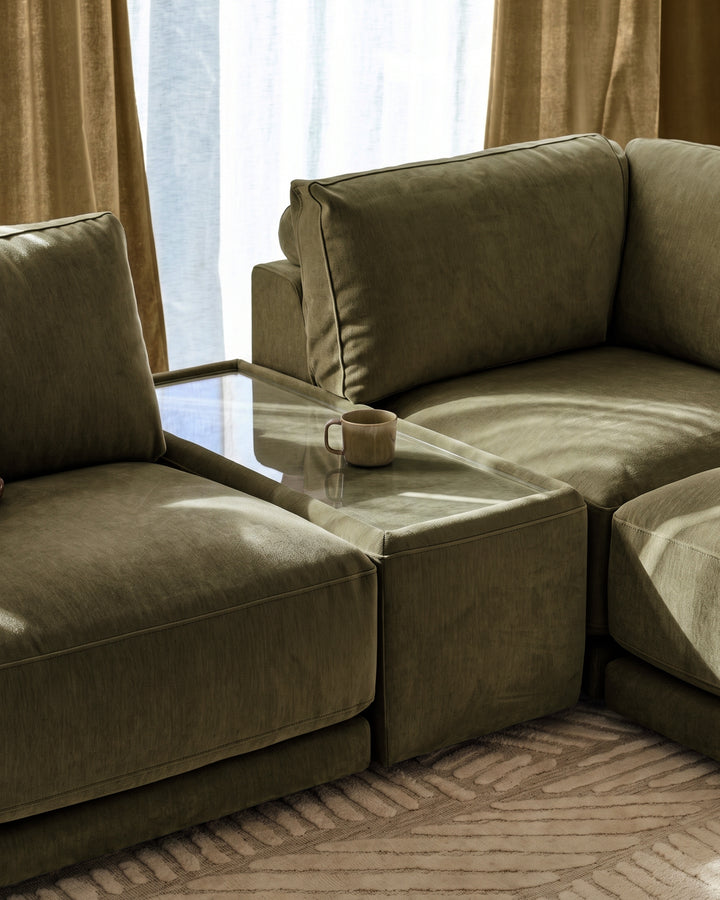 Eden side table module in dark green fabric with glass top, positioned between matching upholstered seating in modern living room setting.