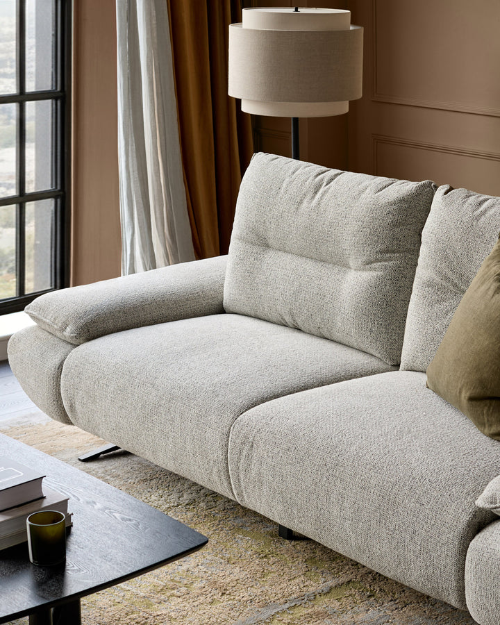 Elio 3-seater sofa in grey fabric, shown at an angle displaying curved armrests and quilted cushions in a modern living room.