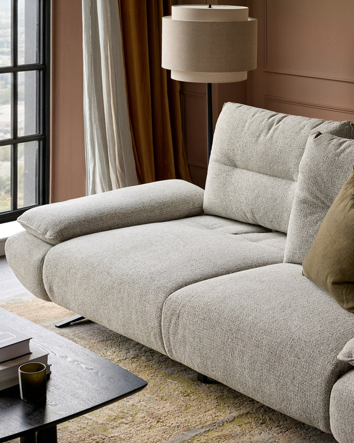 Elio 3-seater sofa in grey fabric with curved, puffy design and padded armrests, positioned in a modern living room setting.