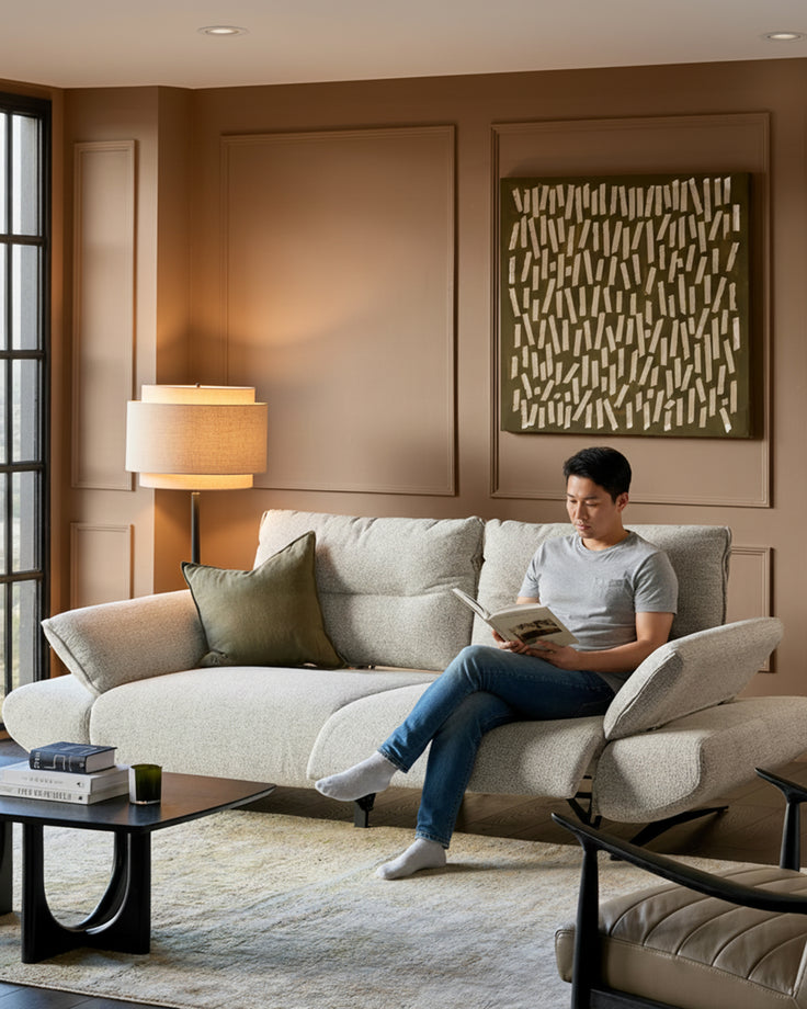 Elio 3-seater sofa in grey fabric with curved design, shown in warm living room with man reading, paired with black coffee table.