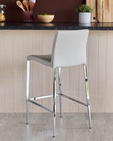 A modern bar stool with a light grey seat and polished chrome frame stands on a light wood floor.