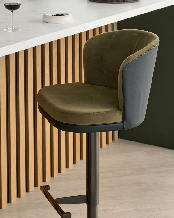 Elodie gas lift bar stool in olive green velvet and black faux leather, shown at kitchen counter with wooden slat detailing.