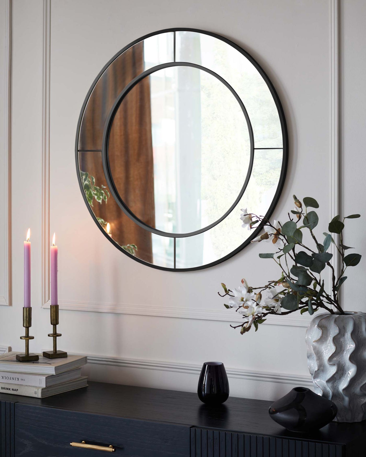 A contemporary dark wood sideboard with a textured front, featuring a large round mirror with a thin black frame hung above it. The sideboard is adorned with decorative elements including a sculptural vase with eucalyptus branches, two pink tapered candles in brass holders, books, and two smaller artistic vases.