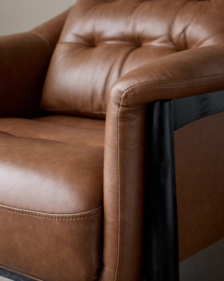 Tan leather accent chair with black wooden frame and armrest, detailed close-up view showing stitching and cushioned seat.