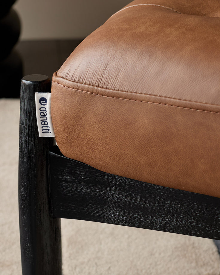 Tan leather accent chair with black wooden frame, showing close-up of curved armrest and brand label detail.