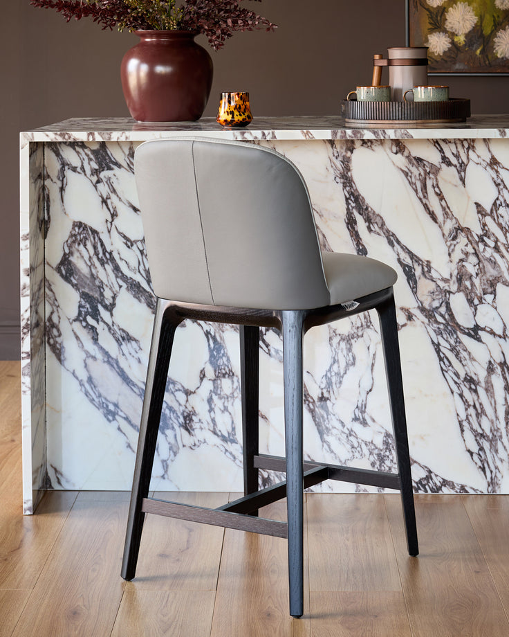 Emerson bar stool in light grey leather with dark grey wooden legs, shown at a marble counter.