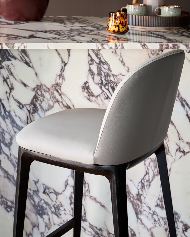 Emerson bar stool in light grey leather with dark grey wooden legs, shown from rear angle at marble table.