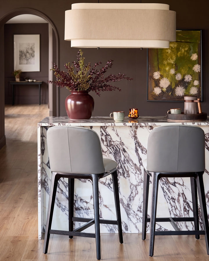 Emerson bar stools in light grey leather with dark grey wooden legs, positioned at a marble counter in a modern dining space.