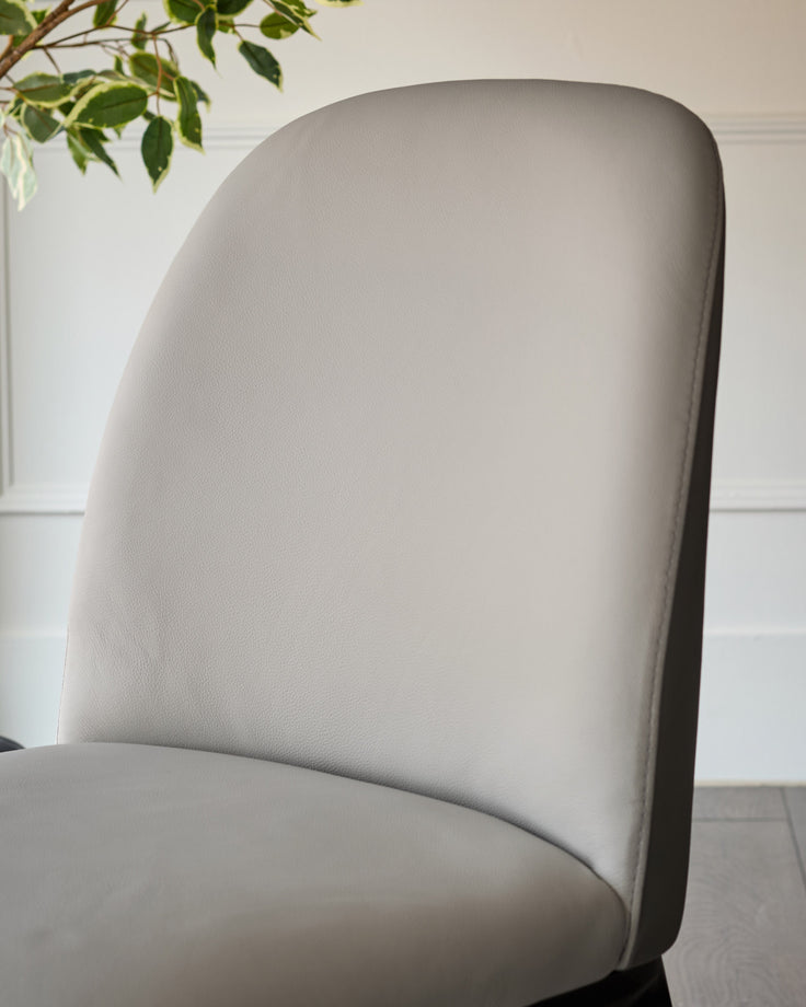 Light grey leather Emerson dining chair with curved backrest, shown from side angle against white wall with greenery.