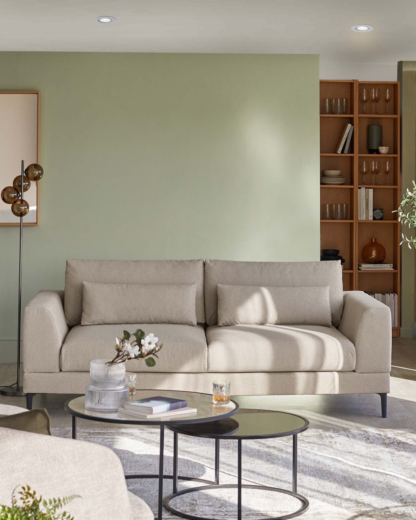 Elegant modern living room featuring a neutral-toned three-seater sofa with clean lines and plush cushions, paired with two round nesting coffee tables with black metal frames and glass tops, set on a textured off-white area rug. A wooden bookshelf with open shelving is in the background, displaying an assortment of decorative objects and books. A stylish floor lamp with a golden finish adds a touch of warmth to the space.